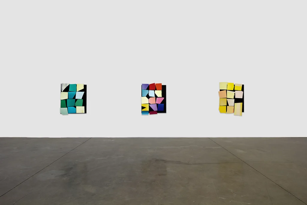 Installation image of An Imprint of the Smallest Gesture that shows three similar works that features a black panel that has yellow, green and multi colors of bright and vivid papers papers collage on the surface