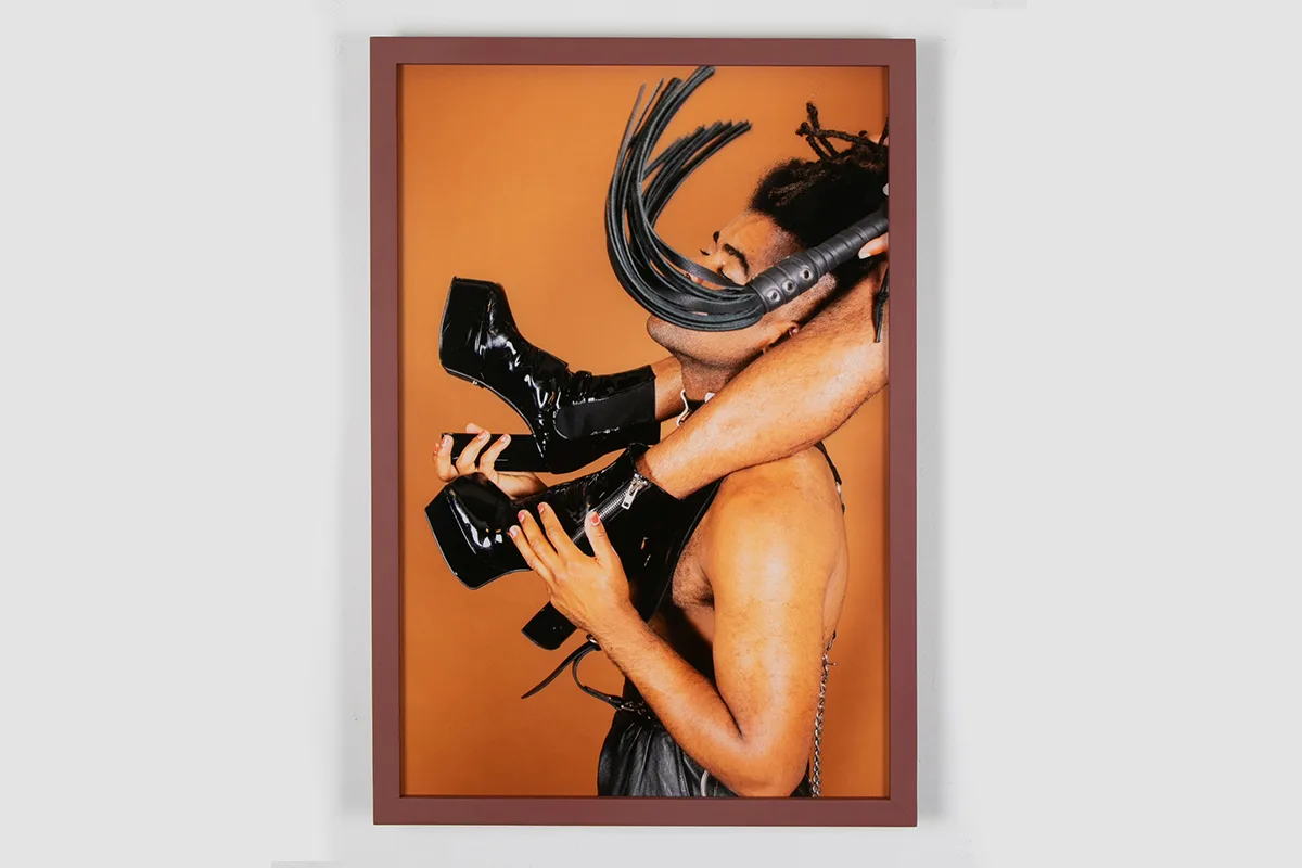 A photo of a two human figures. The first one is standing and wearing a corset and collar while carrying the other person on his upper back. The one on top is wearing a platform shoes and holding a floggers