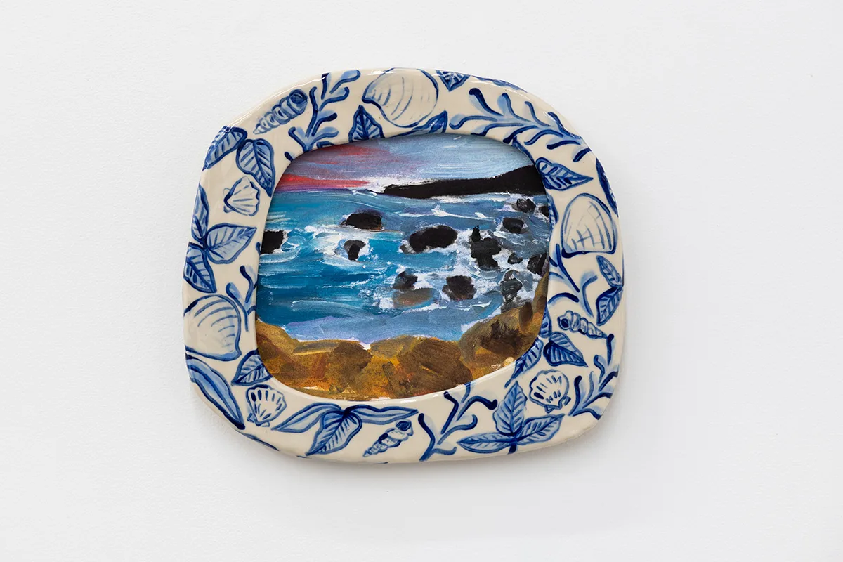 A painted frame of seashells with a painted picture inside of ocean rocks and a sunset over the horizon with mountains in the background by artist Anna Valdez.