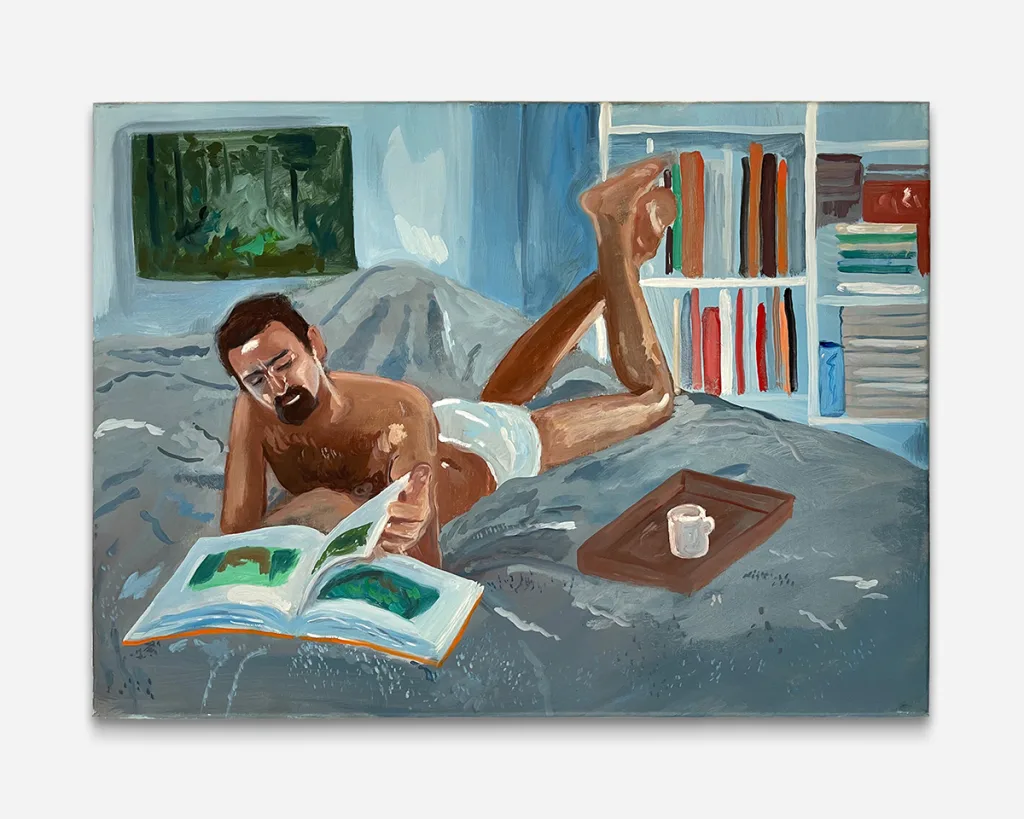 a man in briefs lays with his legs up while reading a book and mug off to the side in bed