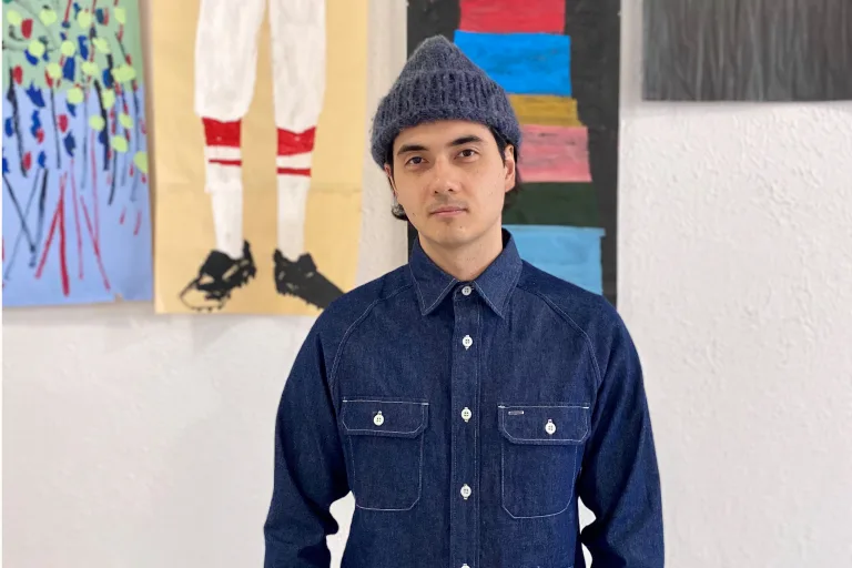 Photo of a man wearing a blue fisherman's cap and denim shirt in front of a wall of paintings