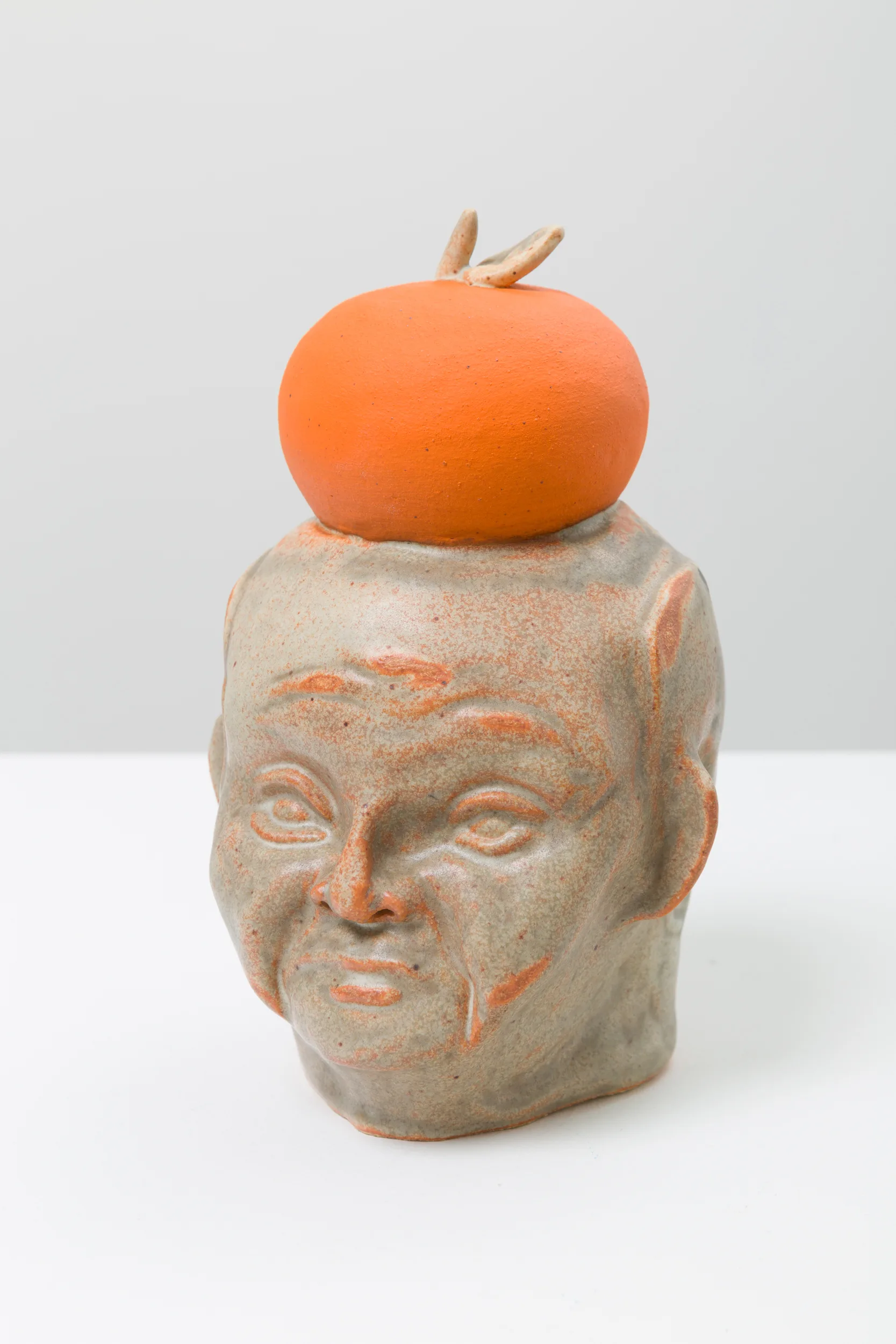 A ceramic bust of a figure with an orange and grey glaze. The figure is angled 3/4 and there is an orange on top of the figure's head