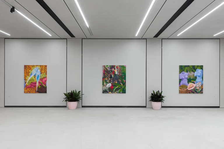 Installation image for Anna Valdez Bird's Eye View shows, about three paintings. First depicts two lovebirds on a branch, second painting are three taxidermized birds and some animal bones. There are striped leaves and flowers surrounding the canvas. And for the third painting featuring two headless woman statues amidst shell, plants, pink flowers, and striped leaves.