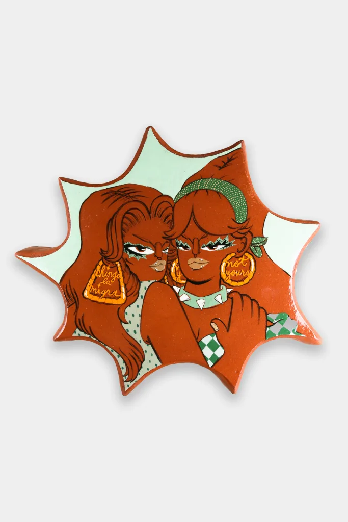 An amoeba shaped glazed ceramic wall-work depicting two women looking forward. The one on the left holds the other in a sheltering manner. They both have confident expressions on their faces.