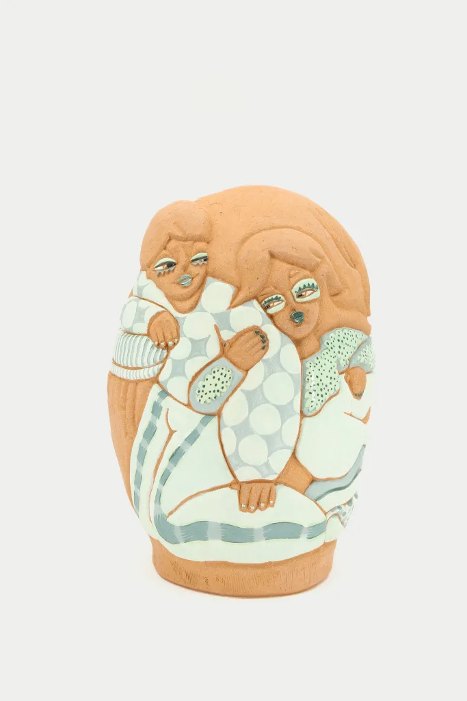 A carved and glazed ceramic vessel depicting two women leaning on each other. They are wearing mint green outfits and the clay body/skin tones are red clay colored. They look towards each other with carefree and comfortable expressions.