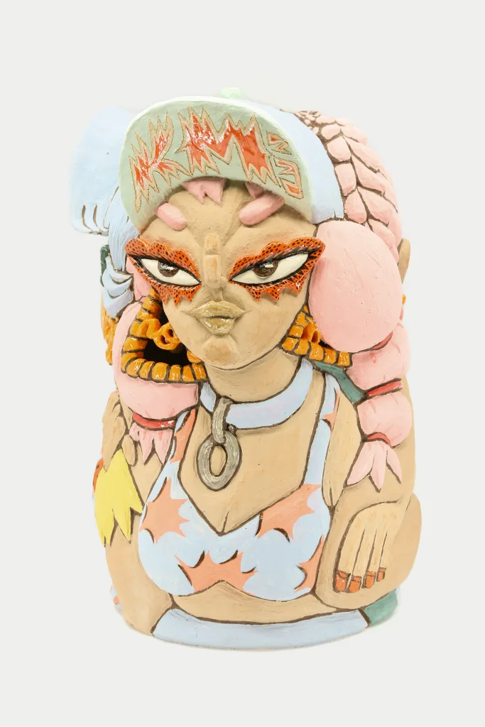 A carved and glazed ceramic vessel depicting three women leaning on each-other. The one in front has fiery red eye shadow, a hat with the brim folded reading the initials KM, and a light blue choker.