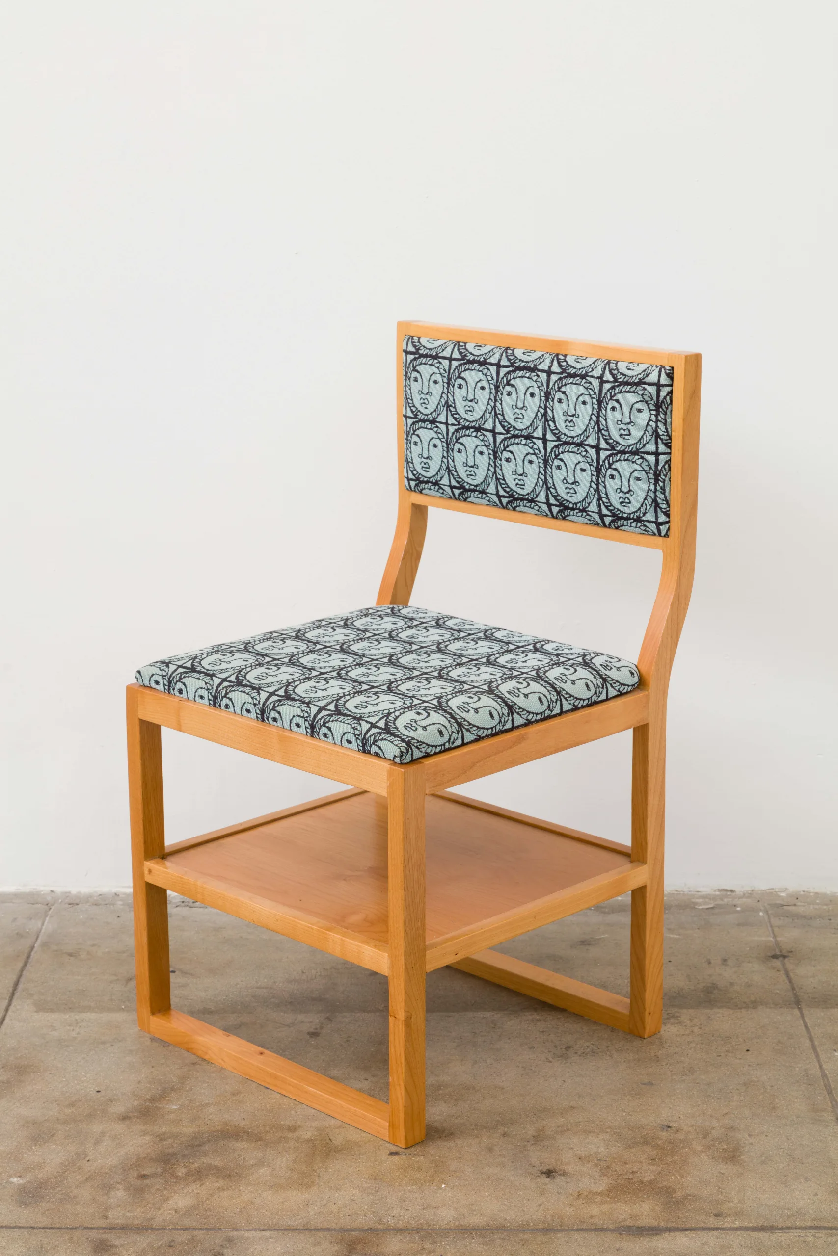 A wooden chair with a shelf under its seat. The seat and back rest cushions are woven and depict a pattern of black line drawings of a woman's face in front of a blue background.