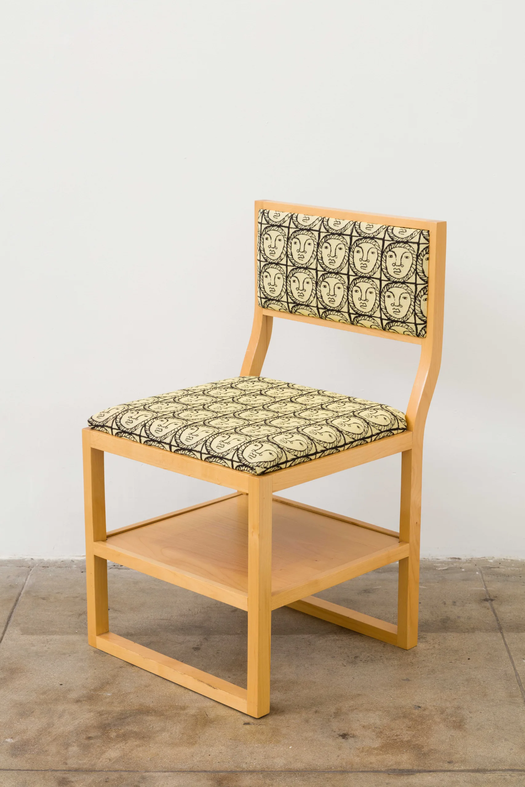 A wooden chair with a shelf under its seat. The seat and back rest cushions are woven and depict a pattern of black line drawings of a woman's face in front of a yellow background.